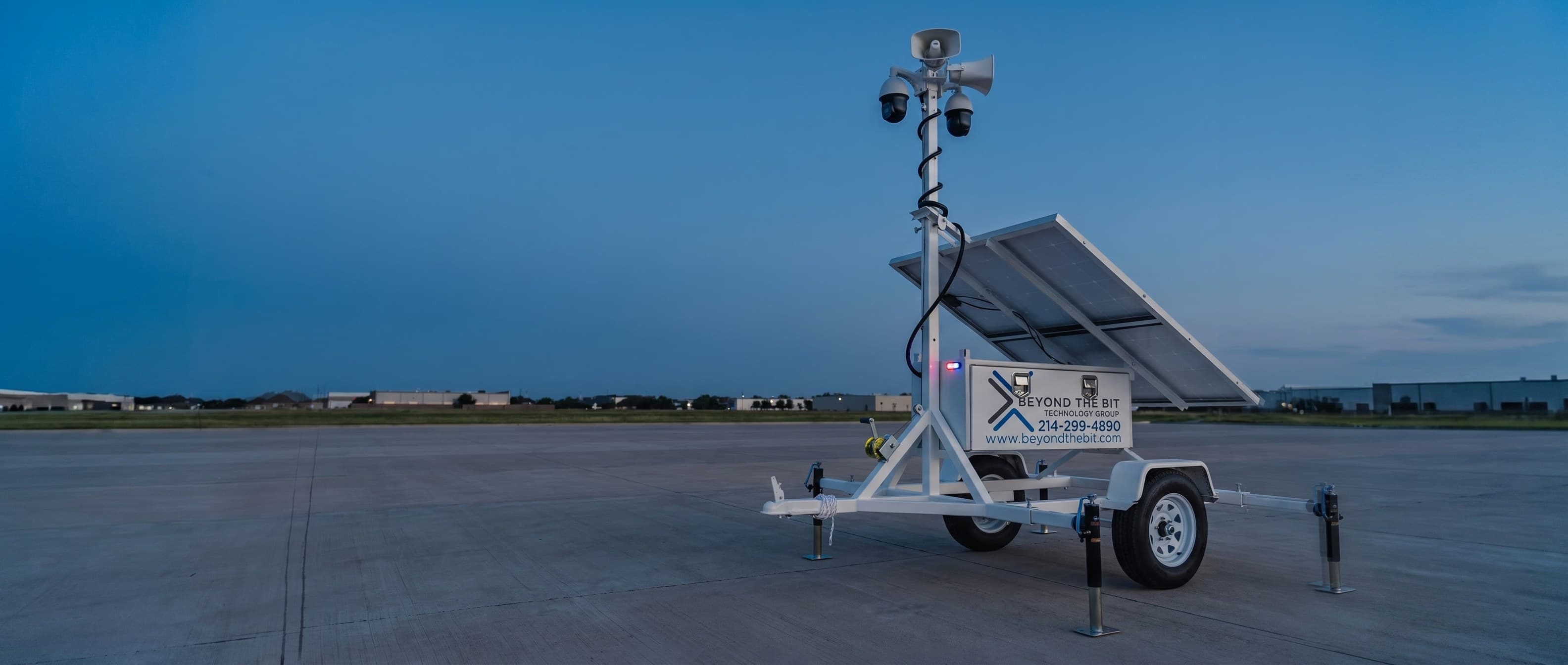 A Beyond the Bit security trailer standing alone on a wide concrete apron at dusk, camera mast fully raised against an open sky.