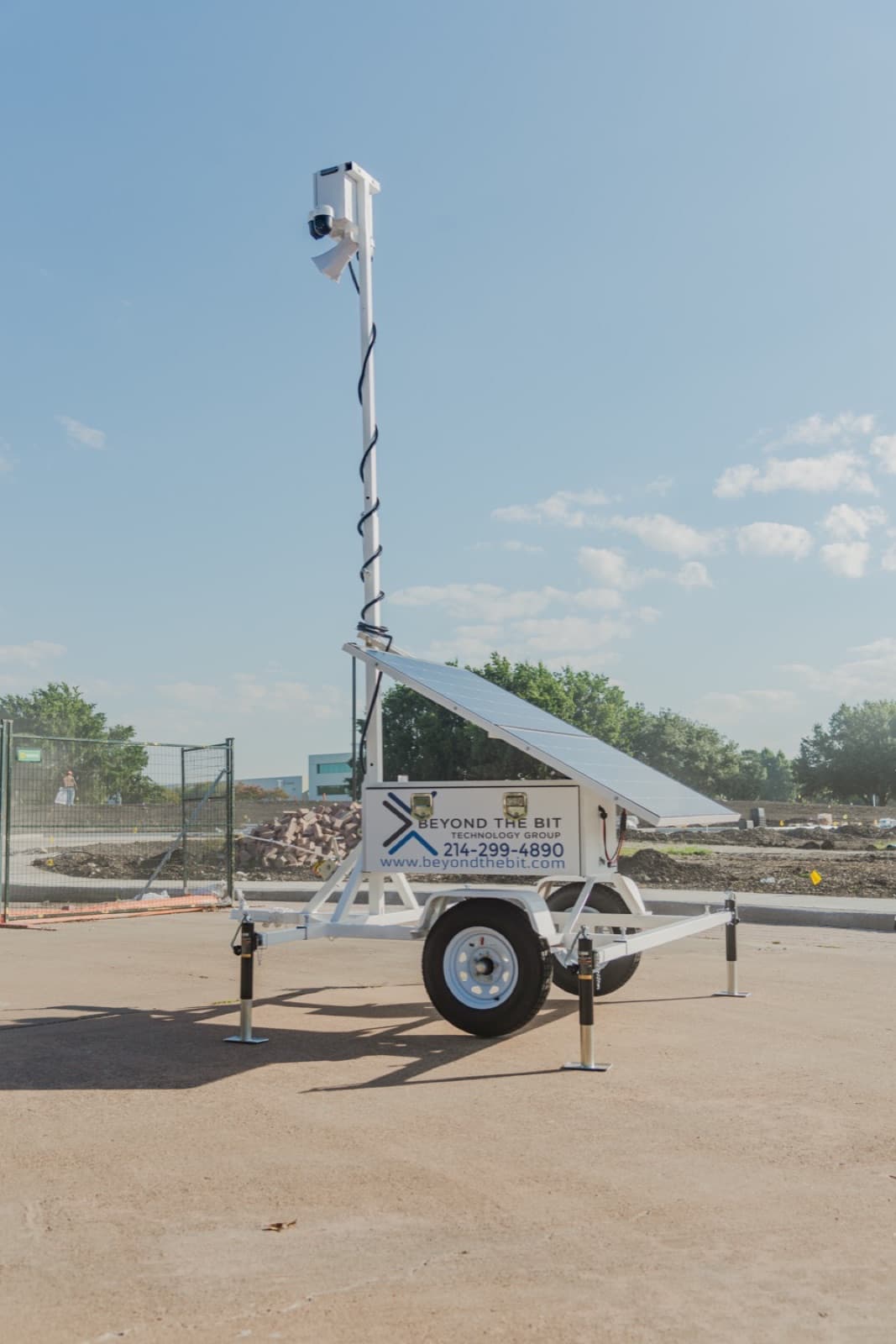 A Beyond the Bit security trailer on a North Texas commercial construction pad, camera mast fully extended, solar array tilted to the sun, four stabilizer jacks down, brick pallets and temporary fencing behind.