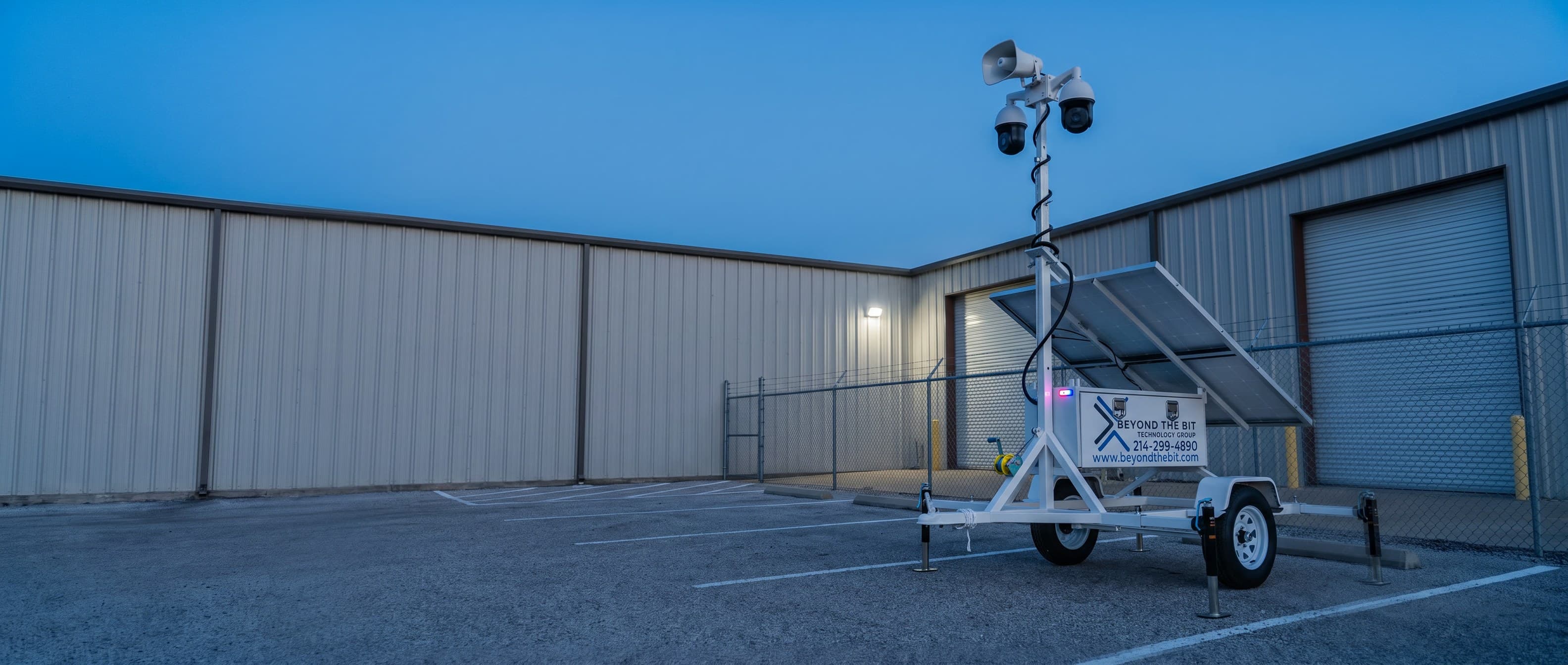 A Beyond the Bit security trailer parked on the gravel hardstand of a fenced equipment yard at blue hour, camera mast raised beside a plain warehouse wall.