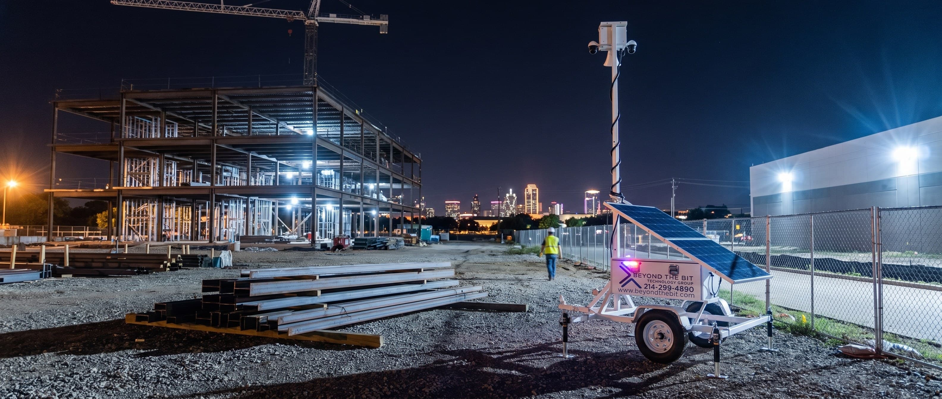 A Beyond the Bit security trailer at night on the gravel pad of an Arlington commercial site, camera mast fully raised beside staged steel beams, a multi-storey steel building frame and tower crane behind.