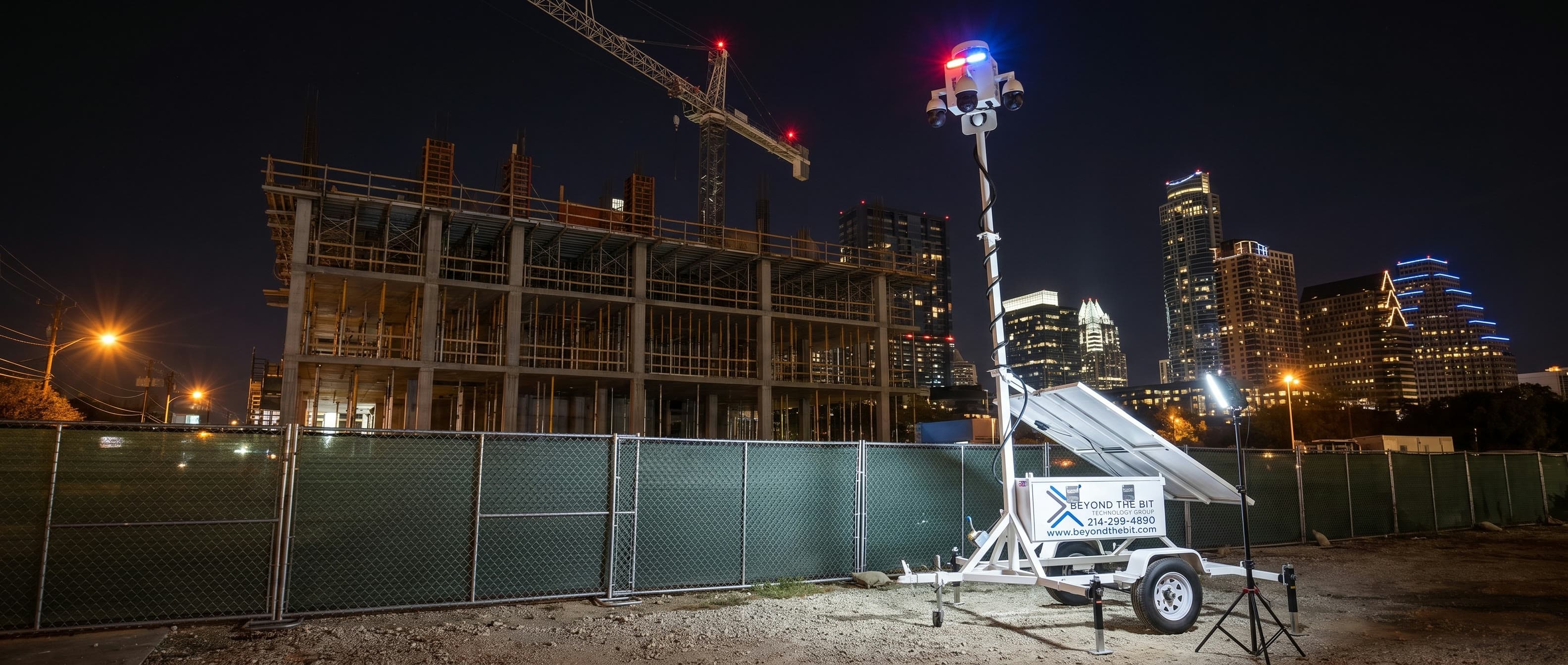 A Beyond the Bit security trailer at night on the gravel pad inside the fence of an Austin mid-rise construction site, camera mast fully raised and its red-and-blue strobe lit, a concrete frame and tower crane behind it and the lit downtown Austin skyline beyond.