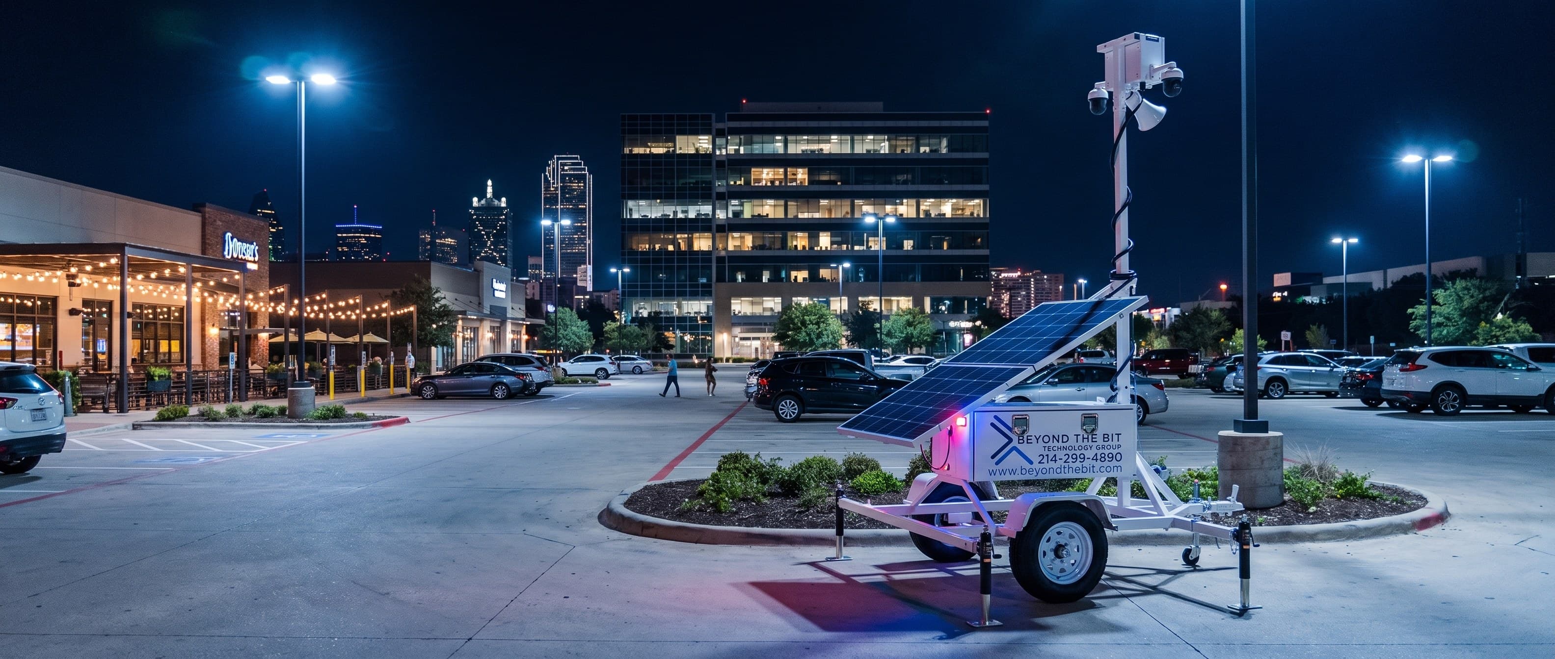 A Beyond the Bit security trailer parked on a lot island of a busy Dallas-area shopping and office plaza at night, its camera mast fully raised, storefronts lit, cars in the rows and people walking to their vehicles.