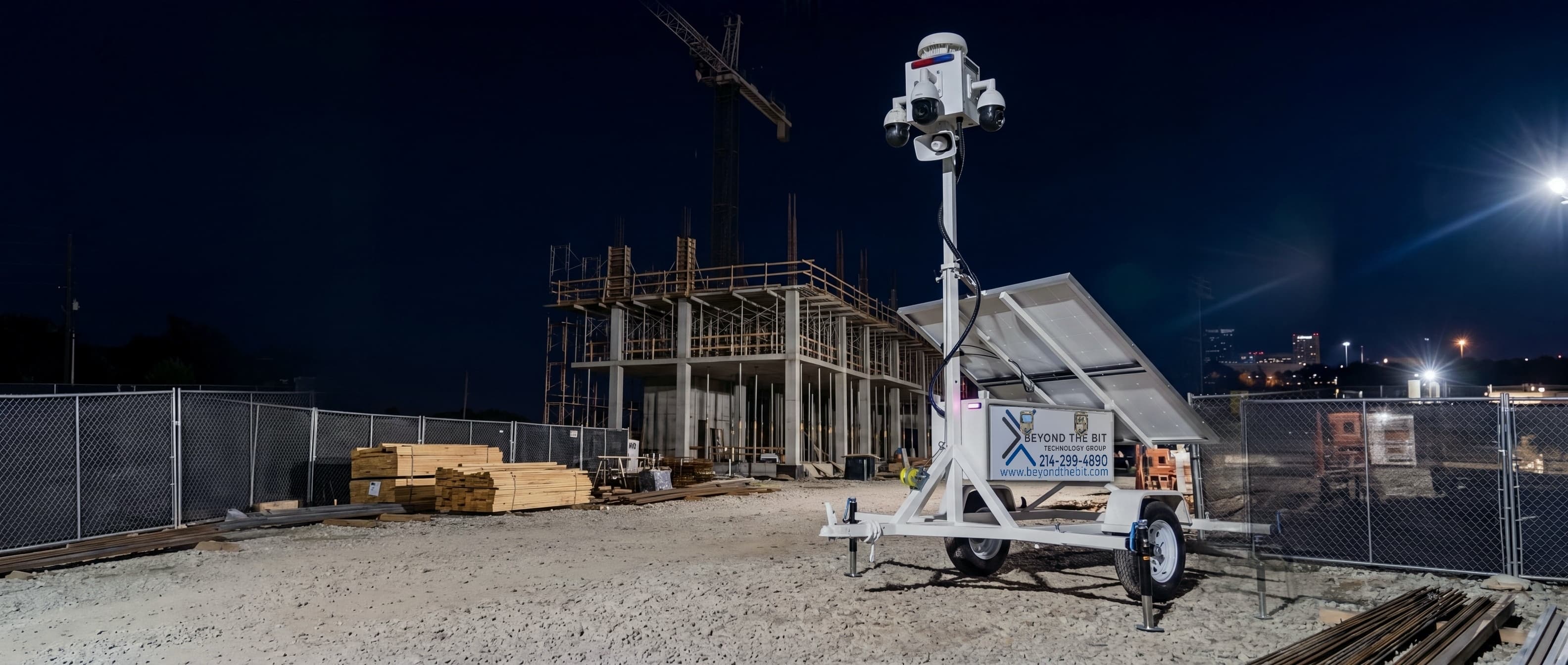 A security trailer at night on an active construction site, camera mast fully raised over staged material inside the perimeter fence.