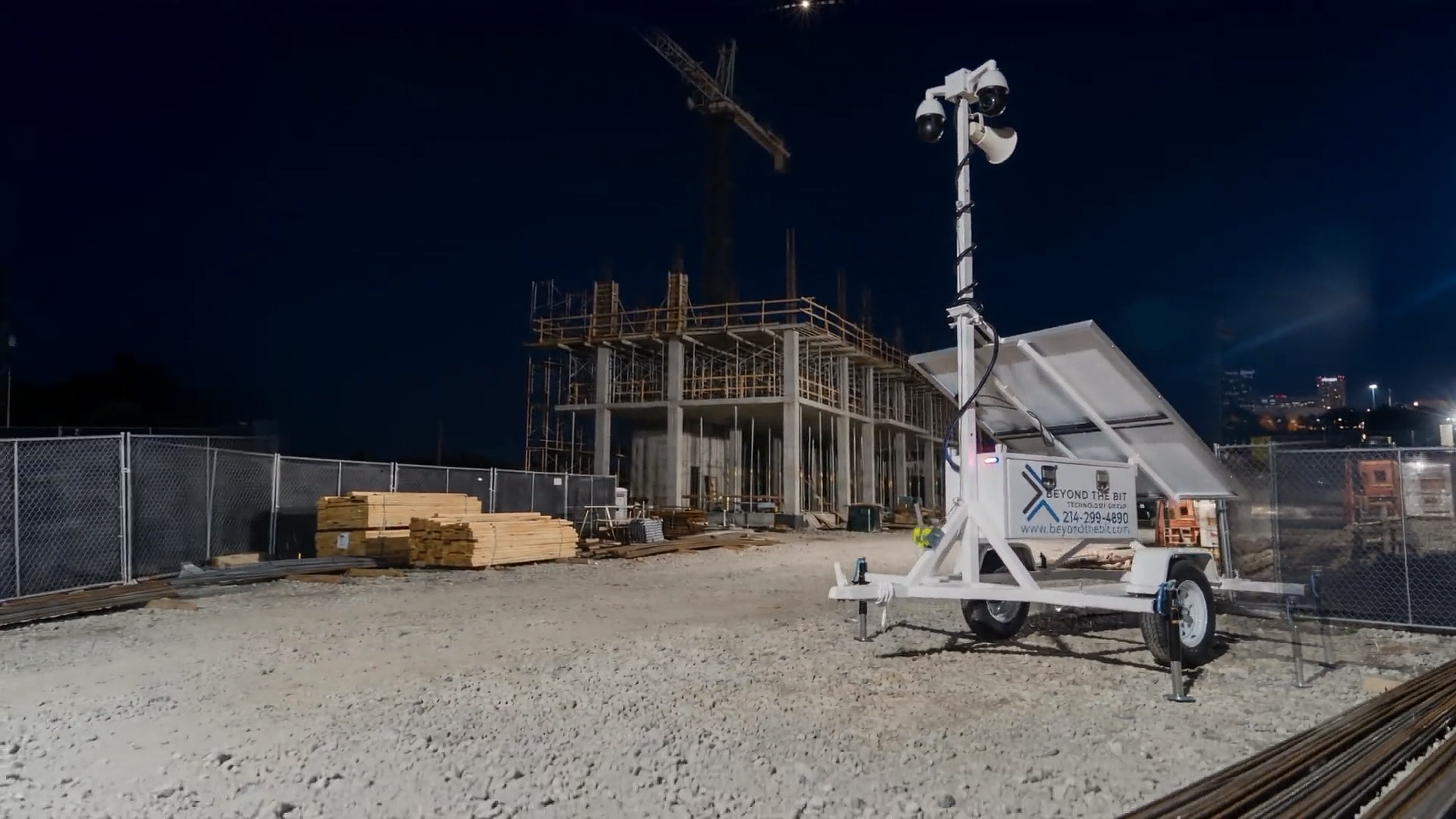 A Beyond the Bit security trailer on a commercial construction site at night, camera mast fully extended above a concrete-frame building under construction, tower crane and stacked lumber behind the perimeter fence.