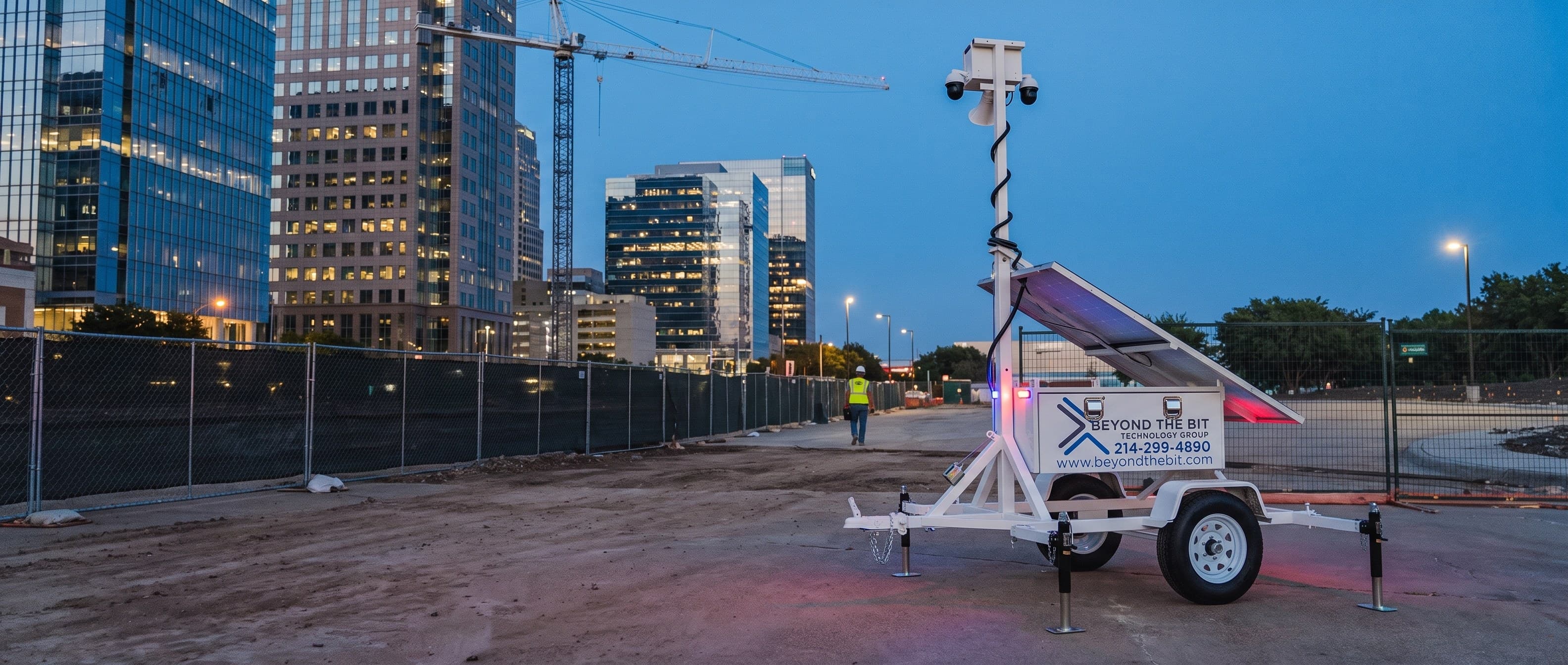 A Beyond the Bit security trailer at blue hour on a fenced infill construction site, its camera mast fully raised, glass office towers and a tower crane standing behind it.