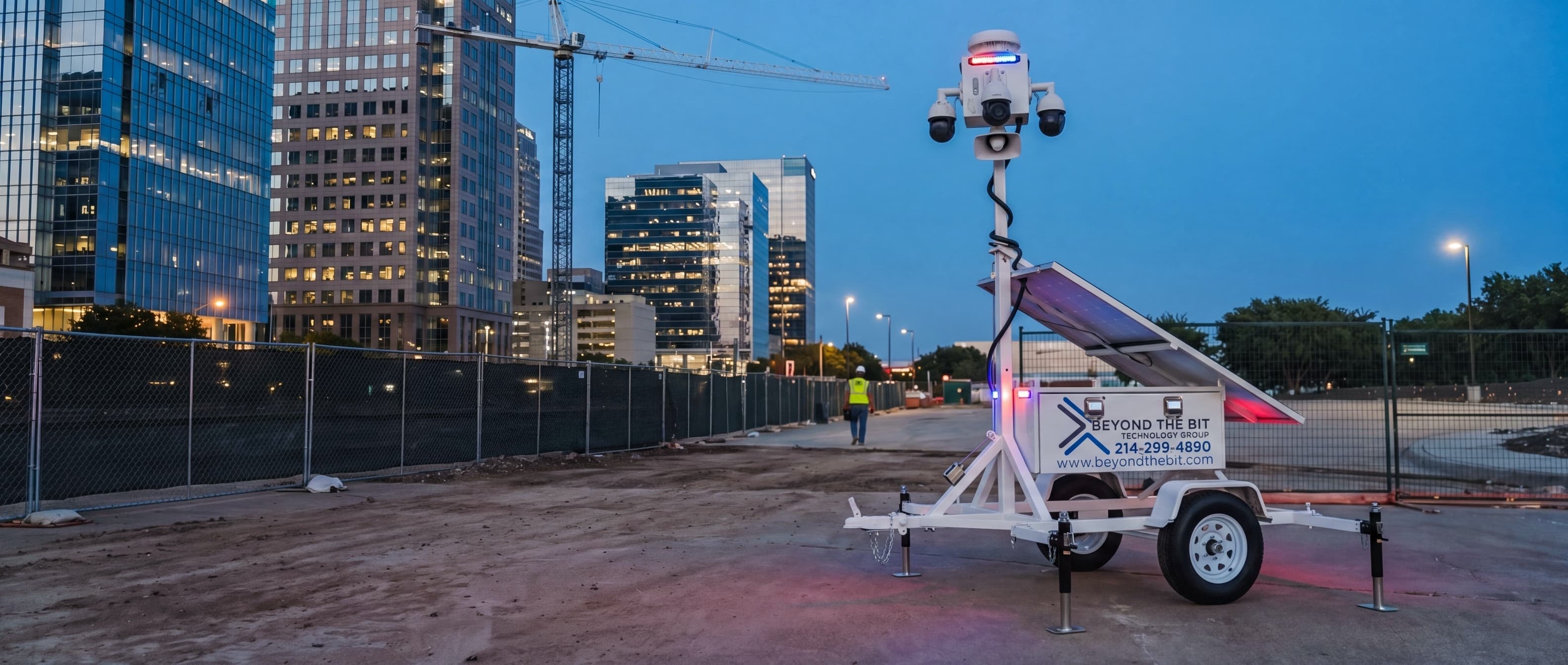 A Beyond the Bit security trailer at blue hour on a fenced infill construction site in the downtown Dallas high-rise core, its camera mast fully raised, glass office towers and a tower crane behind.