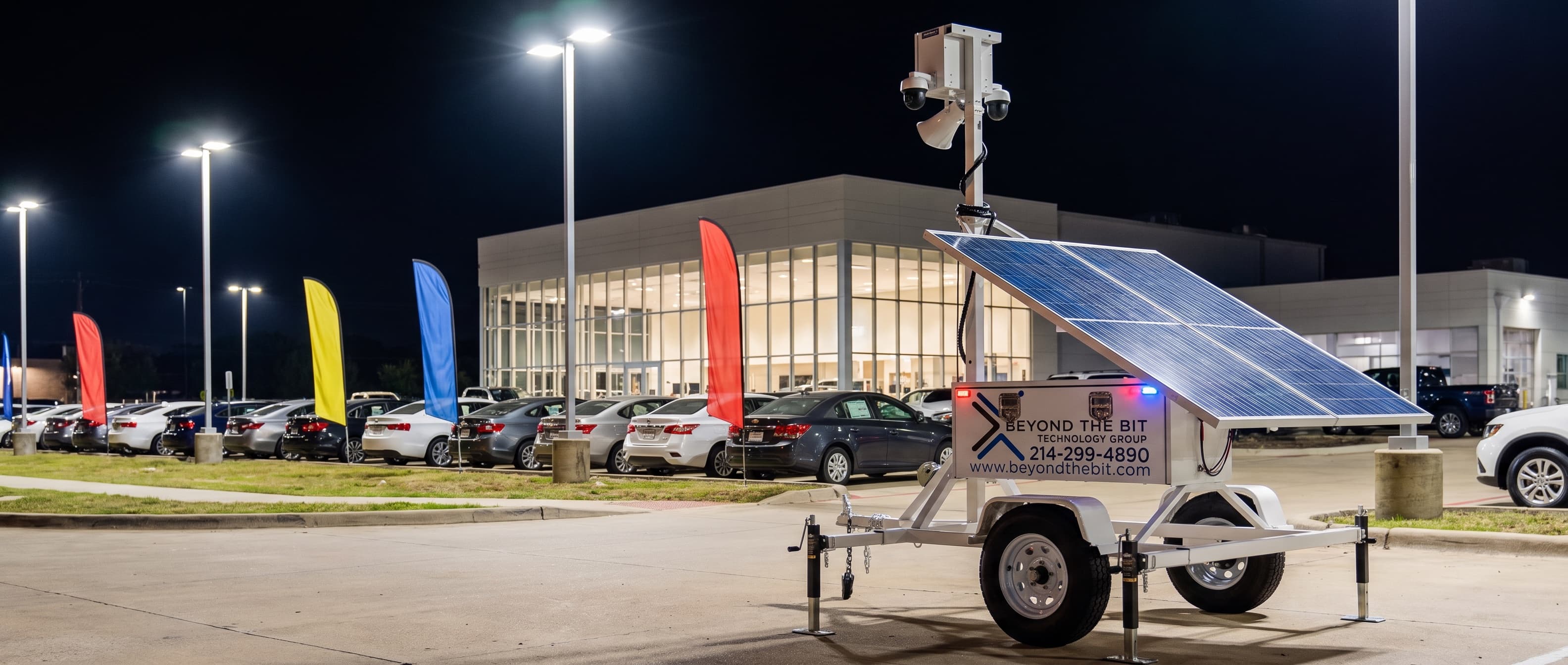 A Beyond the Bit security trailer with its camera mast raised at the street corner of a new-car dealership lot at night, feather flags and a lit glass showroom behind the front row of inventory.