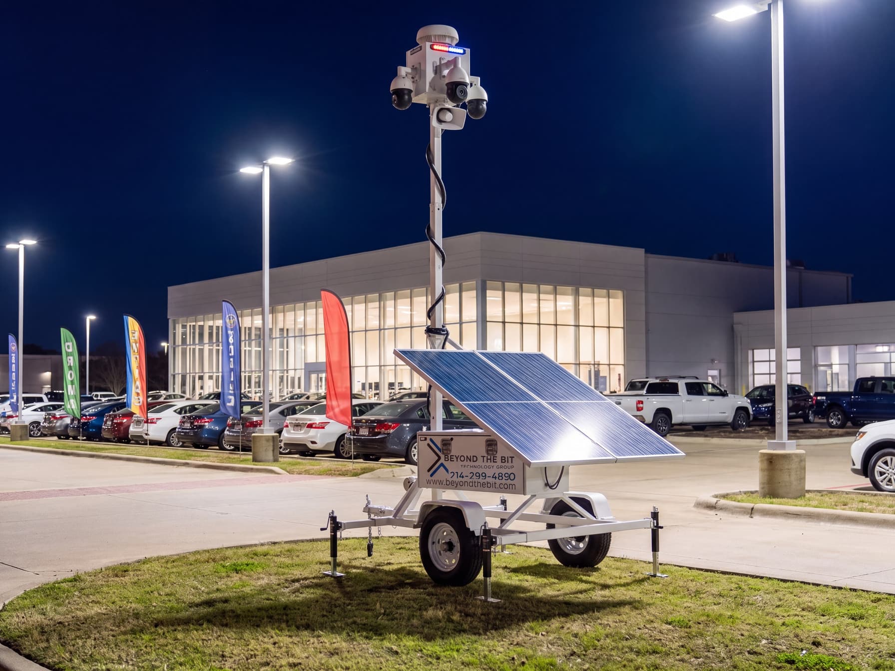 A Beyond the Bit security trailer with its mast raised at the corner of a new-car dealership lot at night, showroom glass glowing behind rows of inventory.