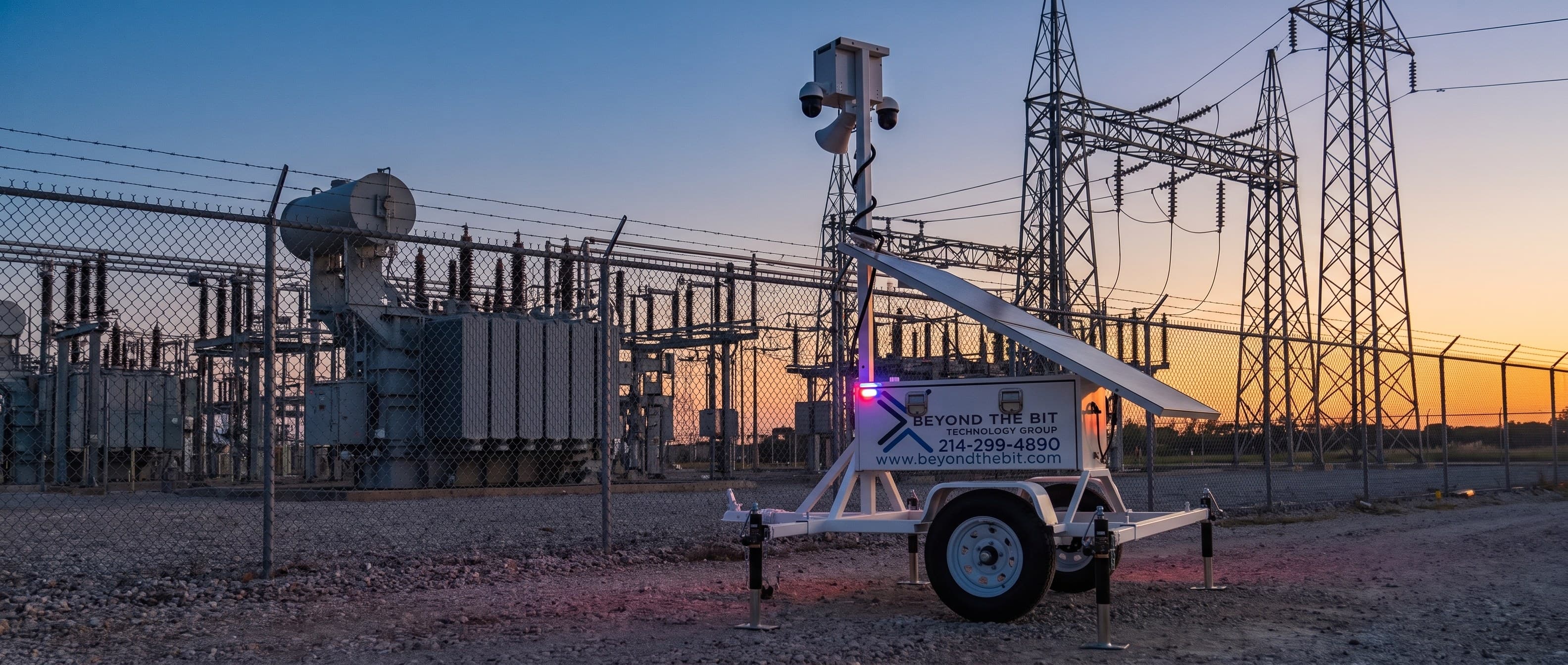 A Beyond the Bit security trailer on the gravel outside a substation fence at sundown, its camera mast fully extended and strobe lit, transformers and lattice steel silhouetted against an orange sky behind the barbed wire.