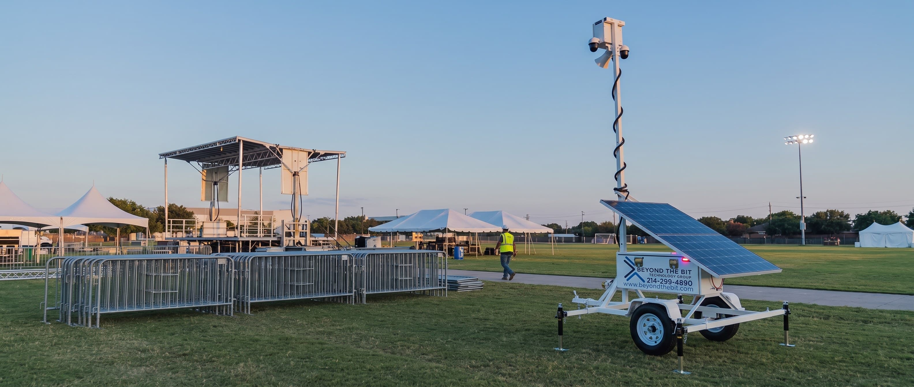 A Beyond the Bit security trailer standing on the grass of municipal event grounds at dusk, camera mast fully raised beside stacked crowd barriers, with a stage frame, event tents and a field light tower behind.