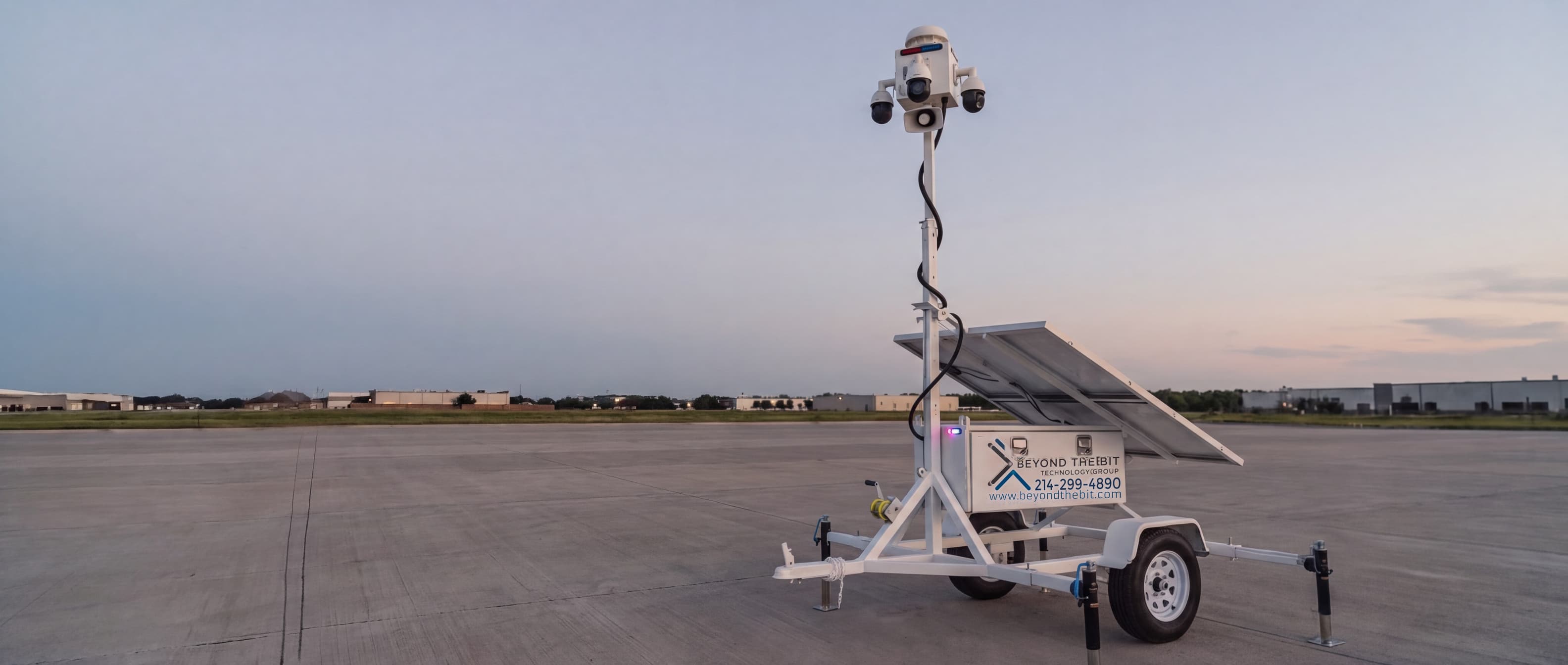 A Beyond the Bit security trailer standing alone on a wide concrete apron at dusk, camera mast fully raised against an open sky.