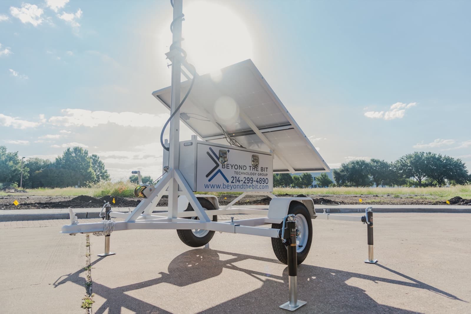 Rear three-quarter view of a Beyond the Bit security trailer, sun flaring over the top edge of the solar panels, the red-and-blue strobe lit on the cabinet corner and the winch visible at the mast base.