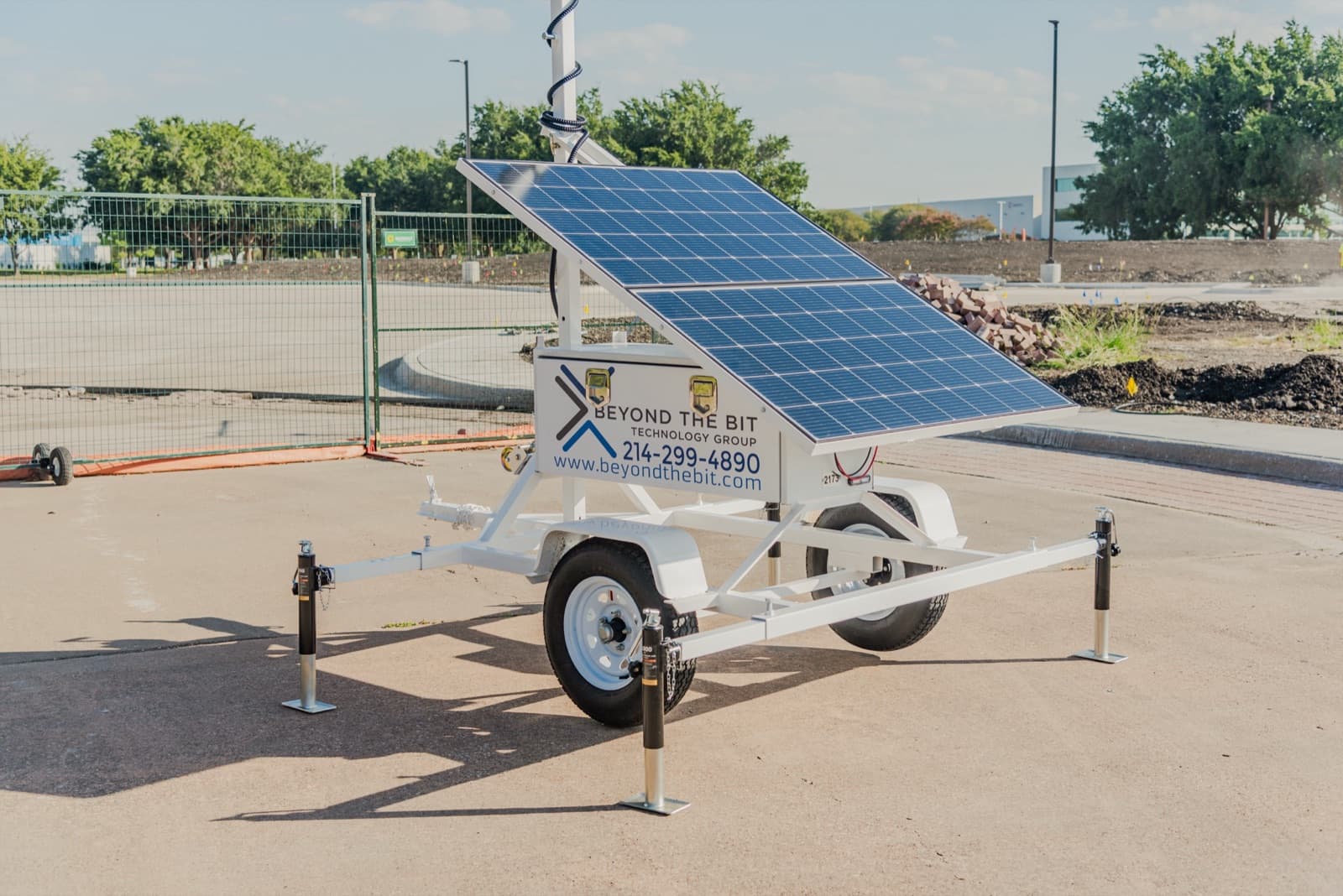 Three-quarter view of a Beyond the Bit security trailer showing the tongue, both wheels, the two-section solar array and the branded equipment cabinet, on a construction pad with green temporary fencing behind.