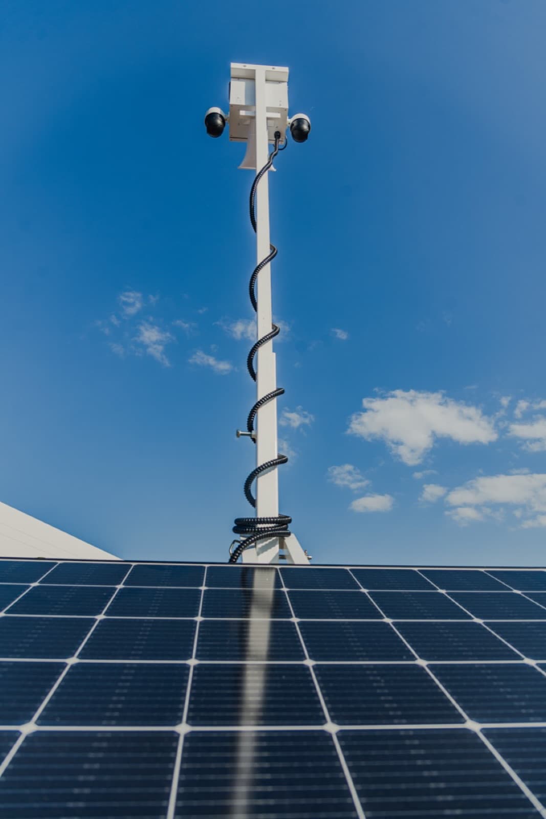 Looking straight up a Beyond the Bit security trailer mast at the head assembly: two dome PTZ cameras on side arms, a white horn speaker beneath, black cable coiled down the mast, solar panels in the foreground.