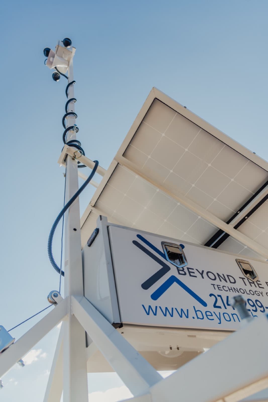 Low angle looking up past the trailer tongue at a Beyond the Bit security trailer mast and camera head against a blue sky, solar panel underside at upper right.