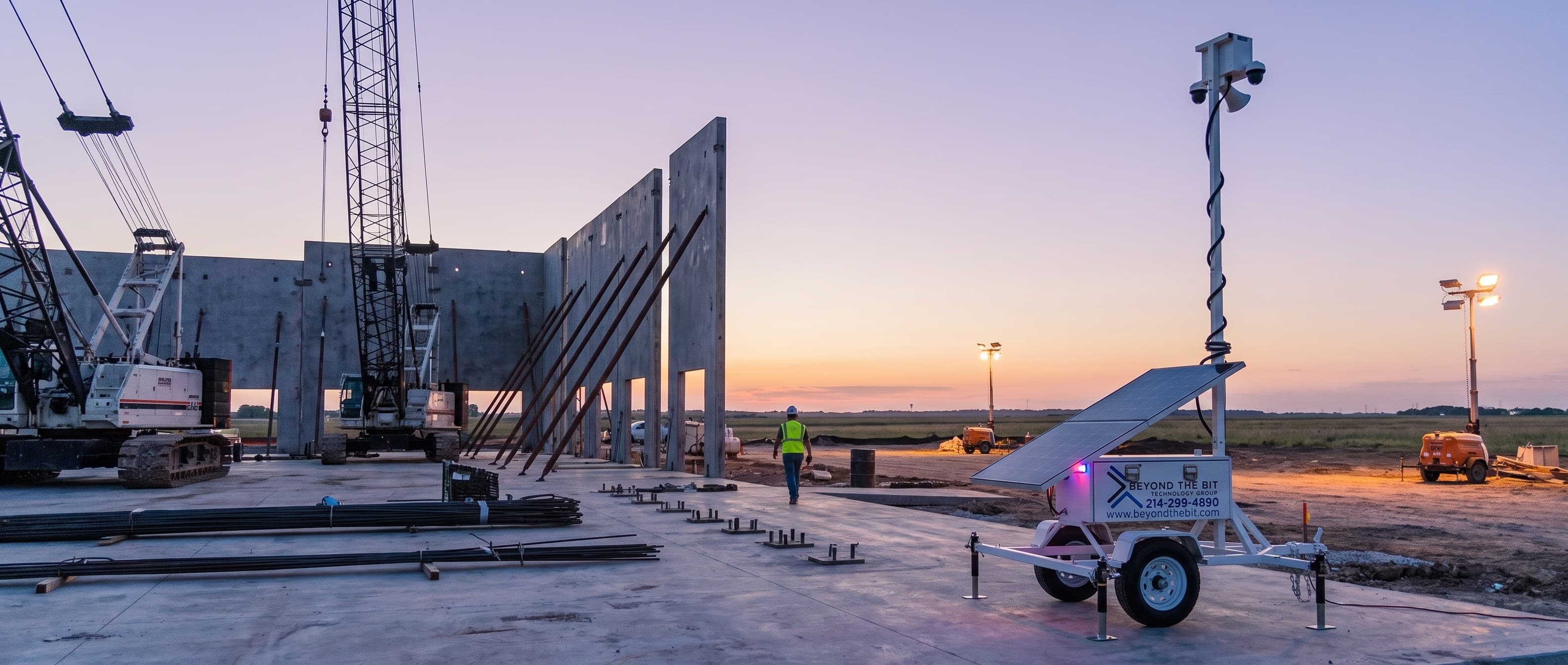 A Beyond the Bit security trailer at dusk on the slab of a Fort Worth-area tilt-wall distribution center build, camera mast fully raised beside freshly braced concrete panels and a crawler crane on a flat prairie horizon.