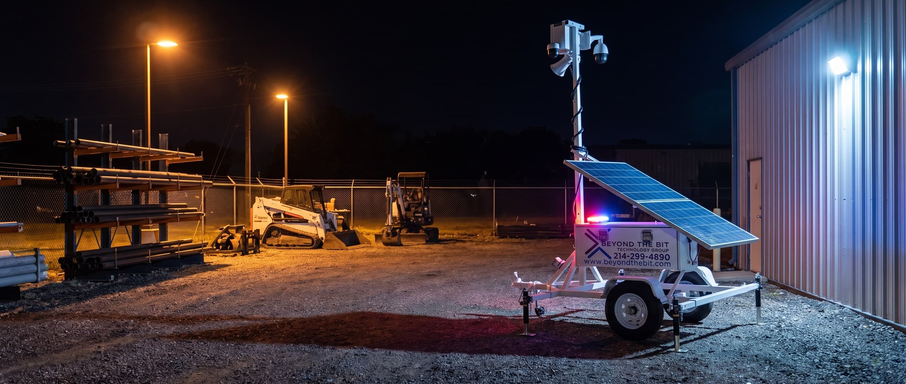 An elevated view of a Beyond the Bit security trailer at night in a Garland industrial equipment yard, camera mast fully extended beside racked steel pipe, a parked skid steer and mini-excavator behind.