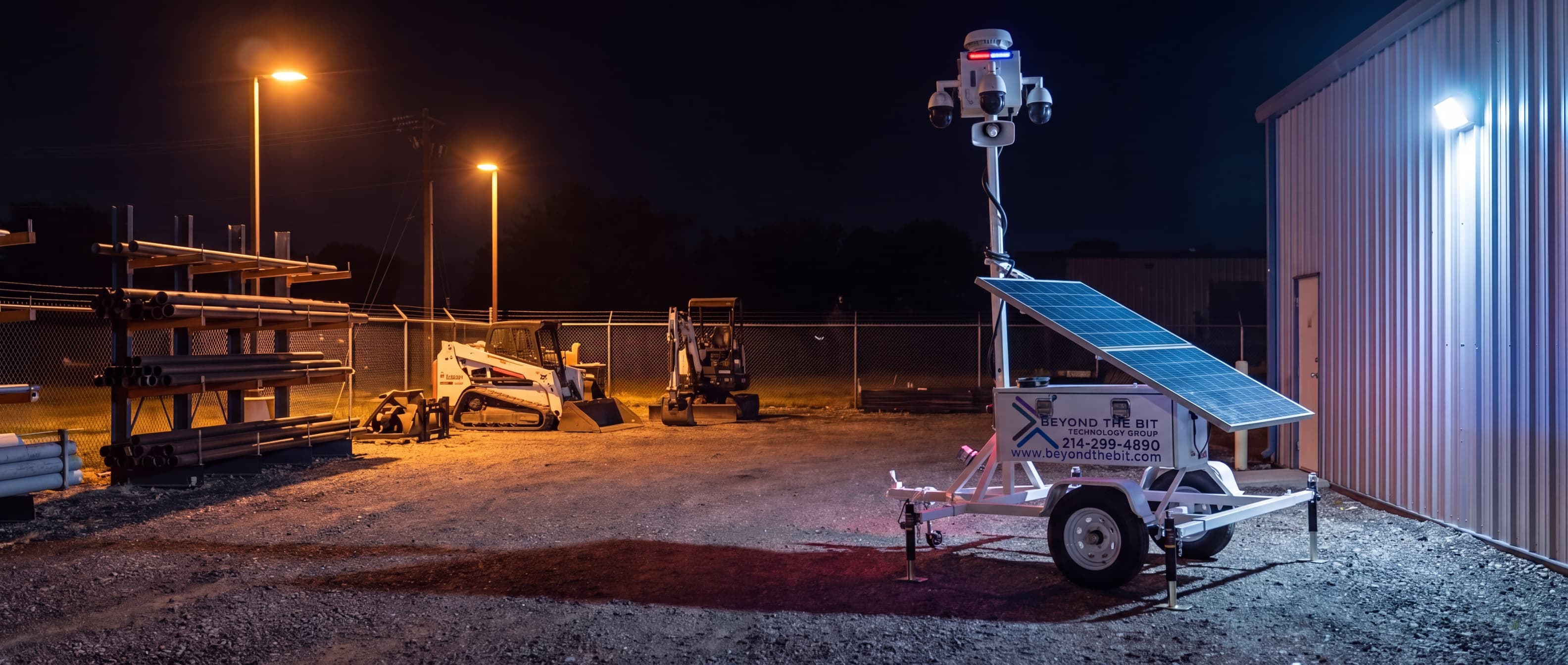 An elevated view of a Beyond the Bit security trailer at night in a Garland industrial equipment yard, camera mast fully extended beside racked steel pipe, a parked skid steer and mini-excavator behind.