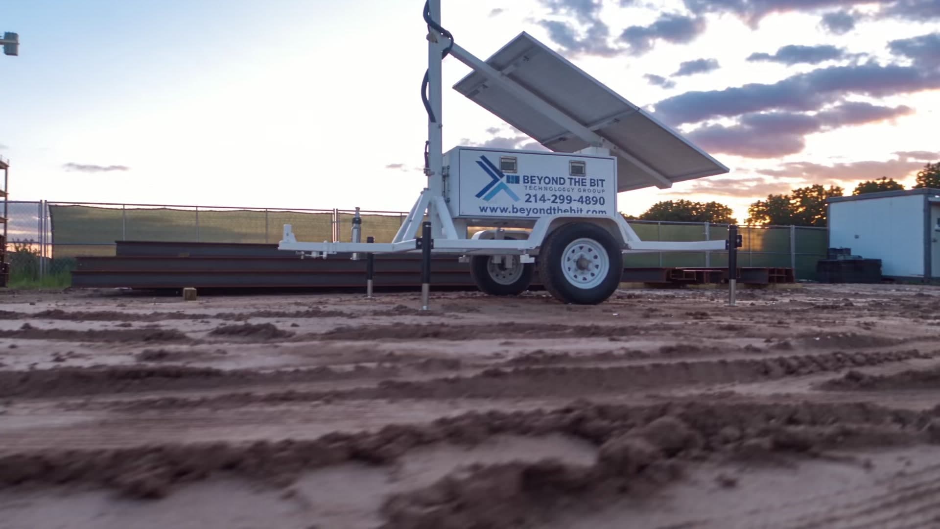 A Beyond the Bit security trailer with solar panel and raised camera mast standing on a dirt laydown yard at dusk, stacked steel beams and a chain-link fence behind it.