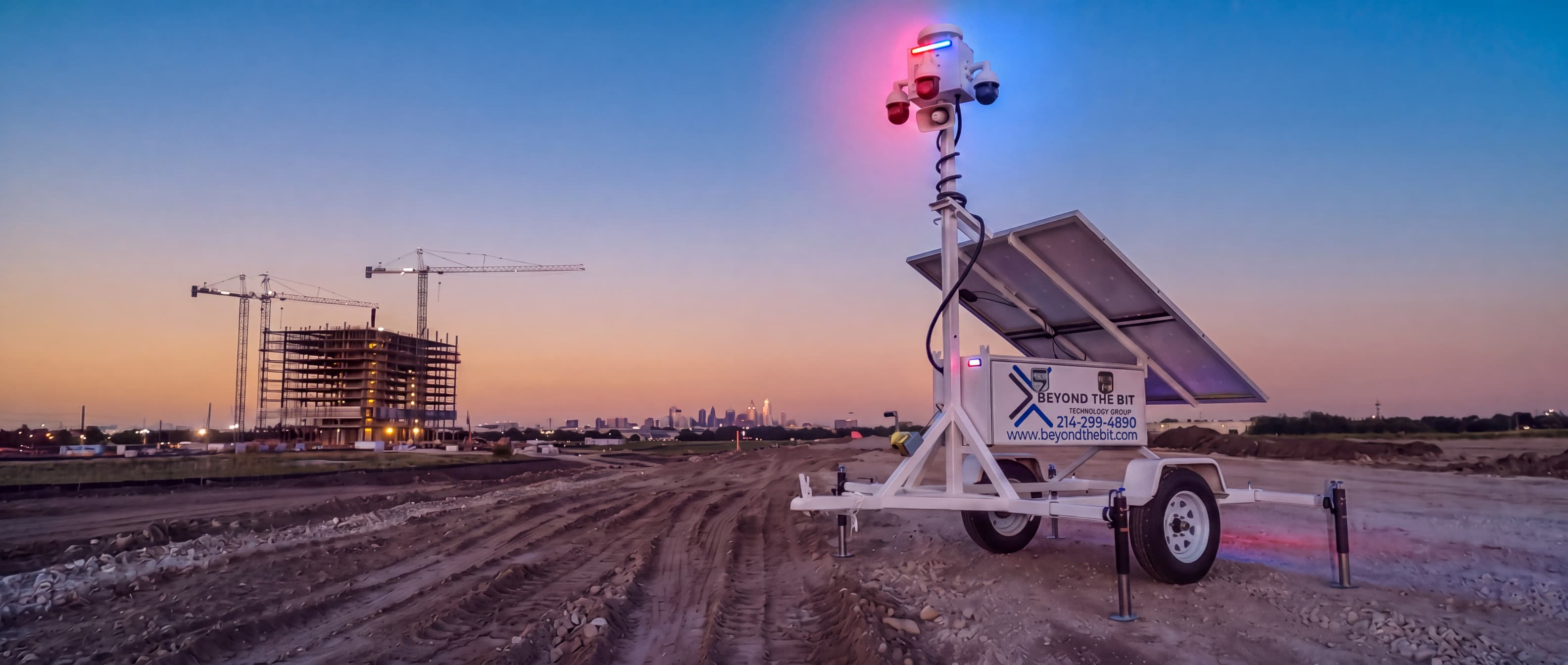 A Beyond the Bit security trailer with raised camera mast standing on a Dallas construction site at dusk, tower cranes and the skyline behind it.