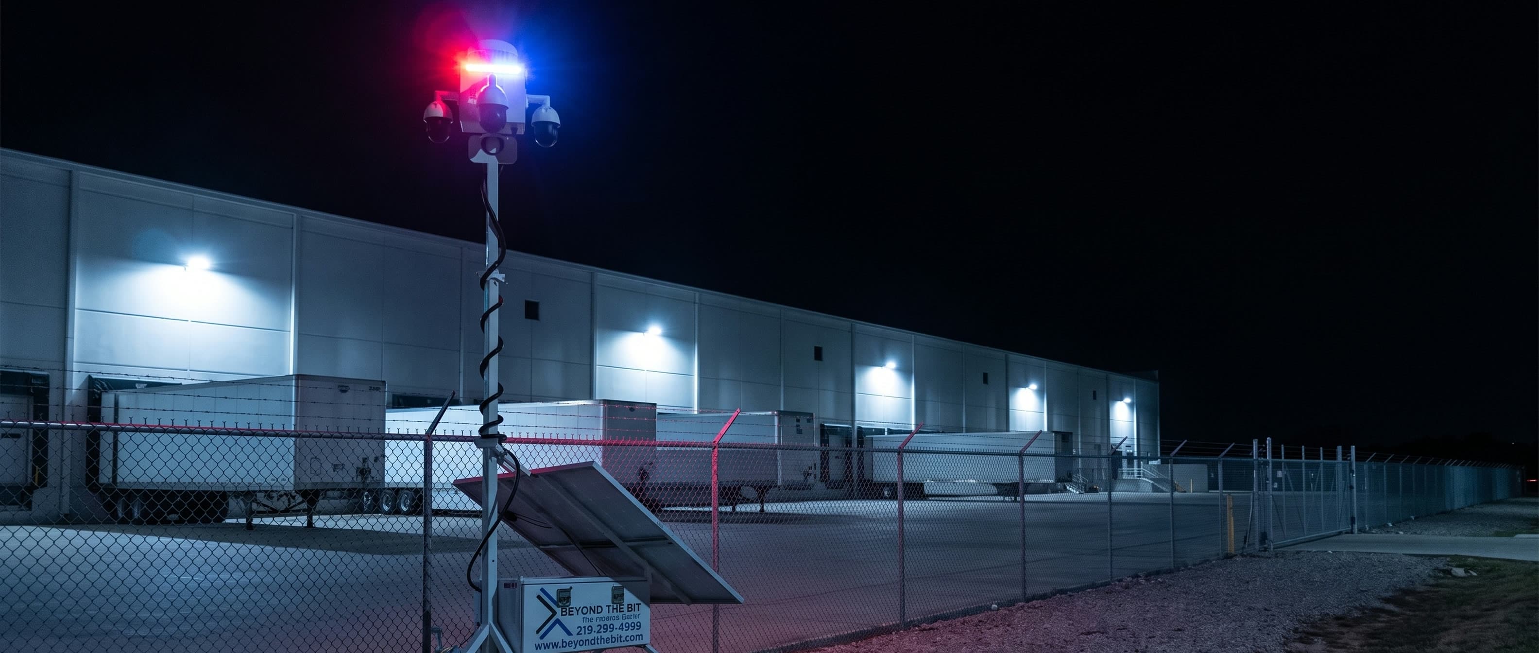 A Beyond the Bit security trailer at night on the gravel outside an Irving distribution-center truck court, parked clear of the gate against the perimeter fence, camera mast fully raised and its red-and-blue strobe lit, trailers standing at the lit dock doors behind it.