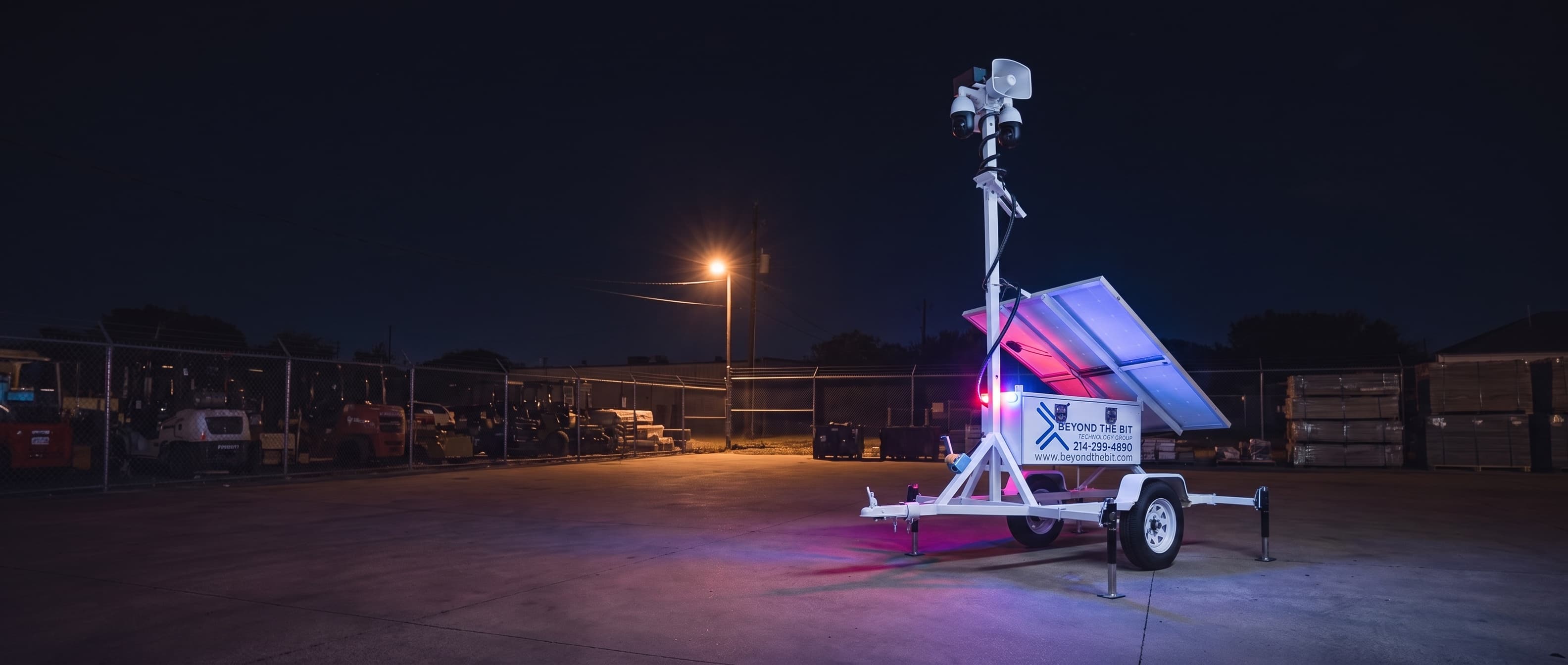 A Beyond the Bit security trailer at night in a fenced equipment yard, the small red-and-blue strobe on the cabinet corner lit and washing the concrete around it, camera mast fully raised.