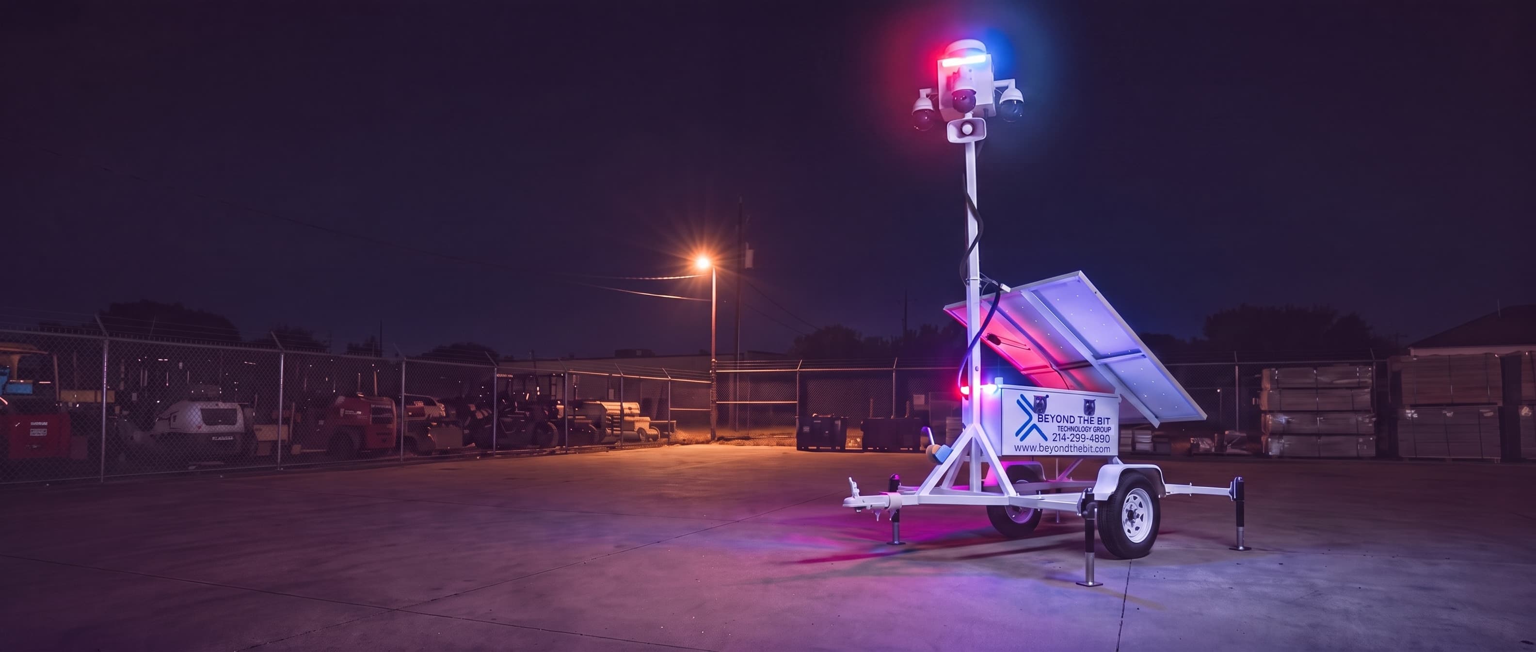 A Beyond the Bit security trailer at night in a fenced equipment yard, the small red-and-blue strobe on the cabinet corner lit and washing the concrete around it, camera mast fully raised.