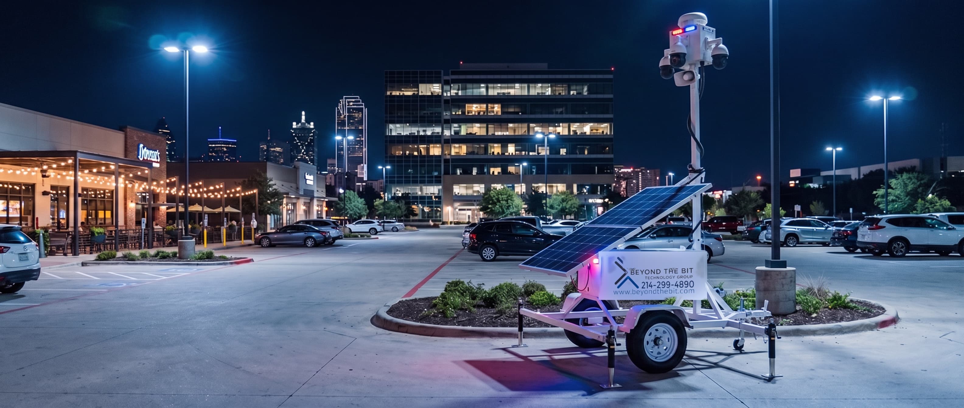 A security trailer at night on a landscaped island of a lit retail and office plaza, camera mast raised over the parking rows after the last customers have gone.