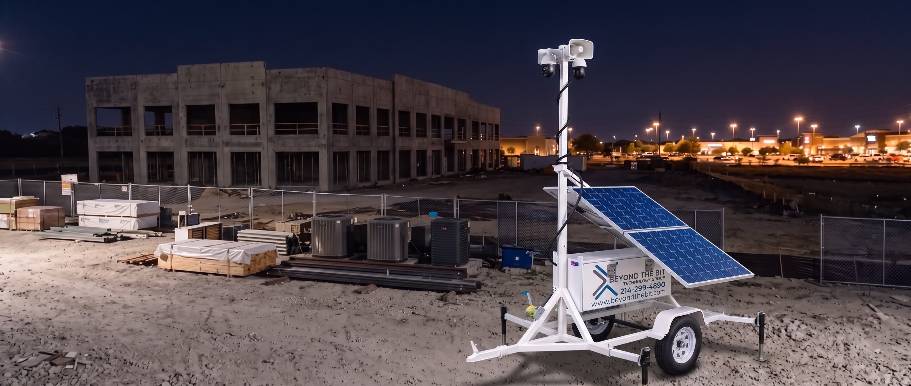 An elevated view of a Beyond the Bit security trailer at night on a Plano office-park construction pad, its camera mast fully extended beside a concrete tilt-wall shell and staged HVAC units, a lit shopping center behind.
