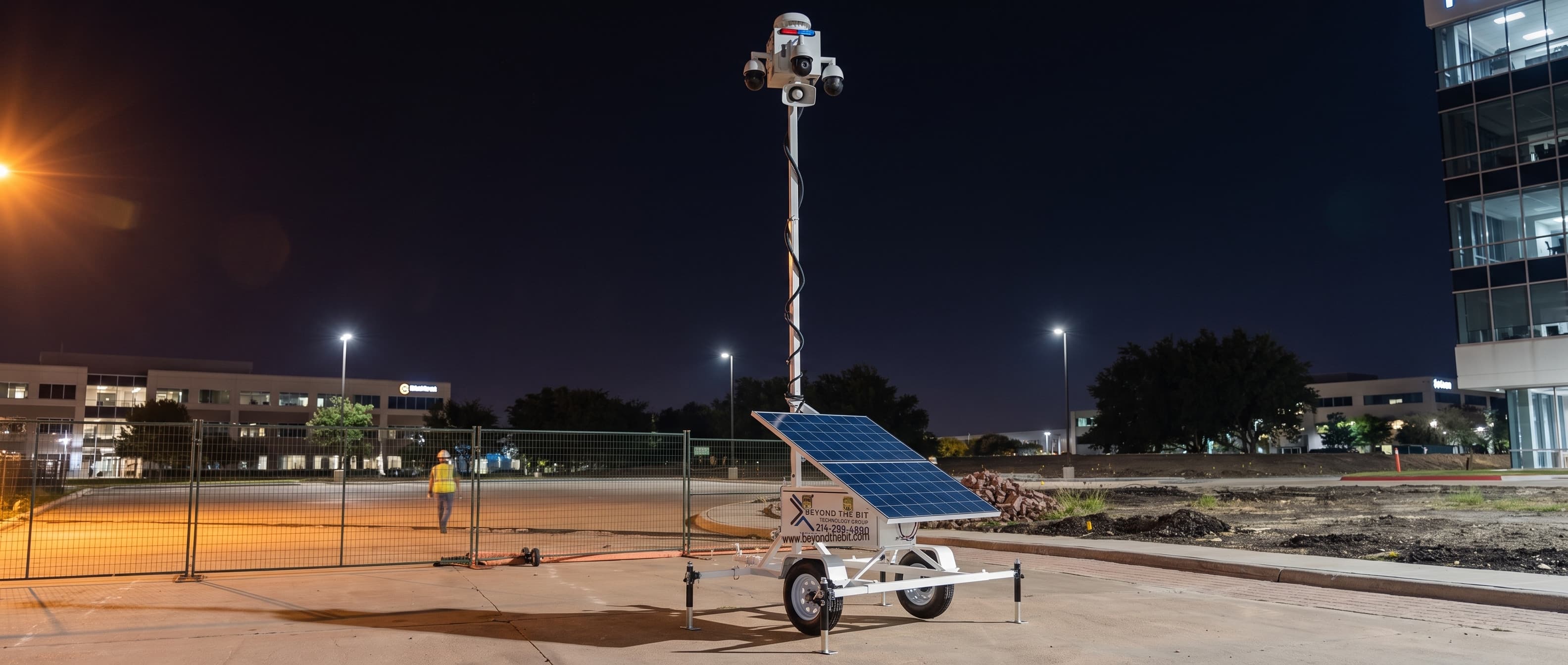 A Beyond the Bit security trailer at night on a fenced Richardson office-corridor pad, camera mast fully raised, rooftop units and coiled conduit staged on pallets, lit glass office floors behind.