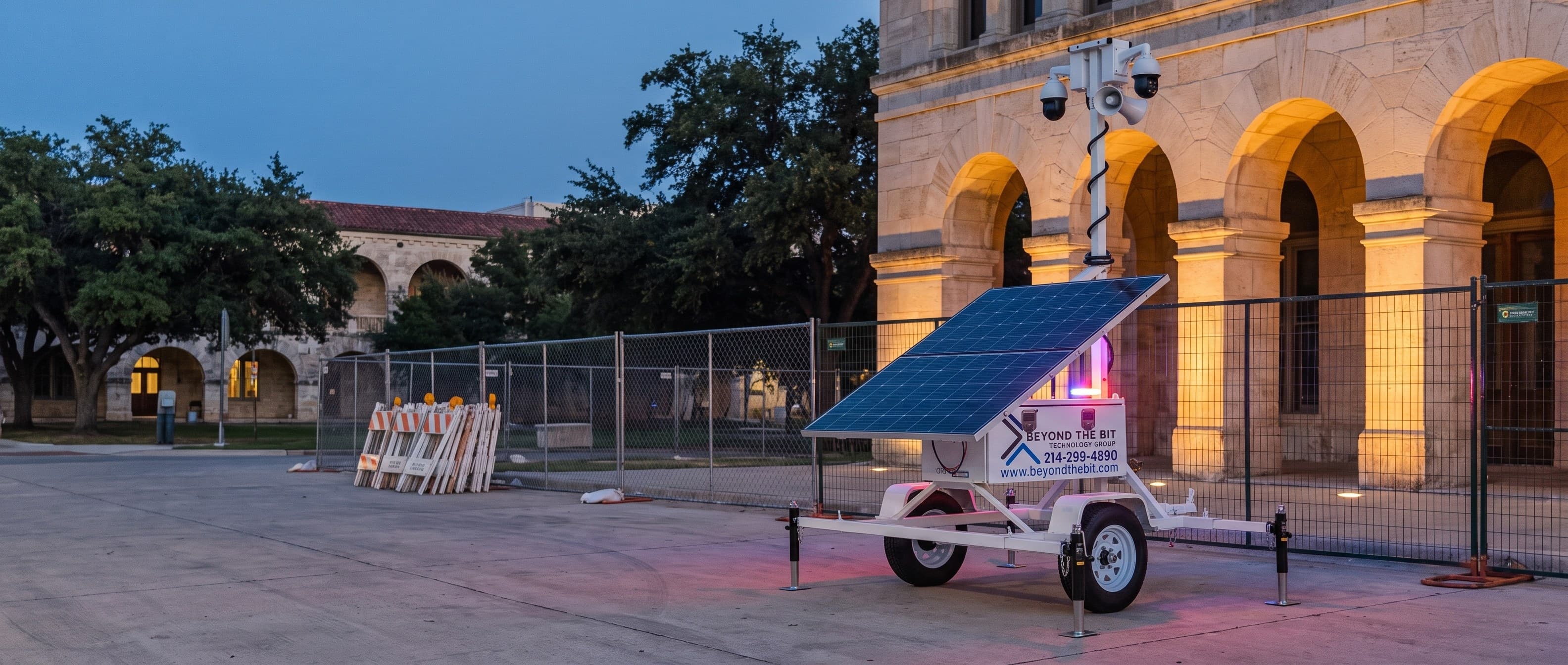 A Beyond the Bit security trailer parked at dusk on the apron outside a fenced civic building under renovation, pale limestone arcades uplit behind it — representative of San Antonio municipal projects.