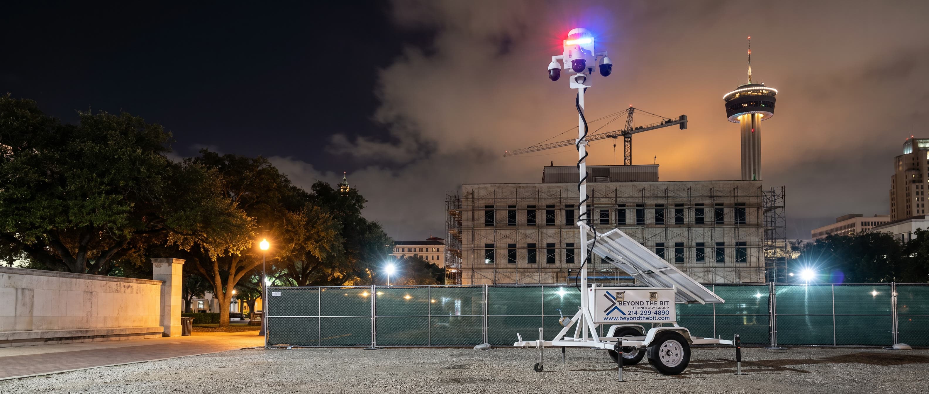 A Beyond the Bit security trailer at night on the gravel staging pad of a fenced San Antonio civic building under renovation, camera mast fully raised and its red-and-blue strobe lit, scaffolding and a tower crane behind it and a lit observation tower on the skyline.
