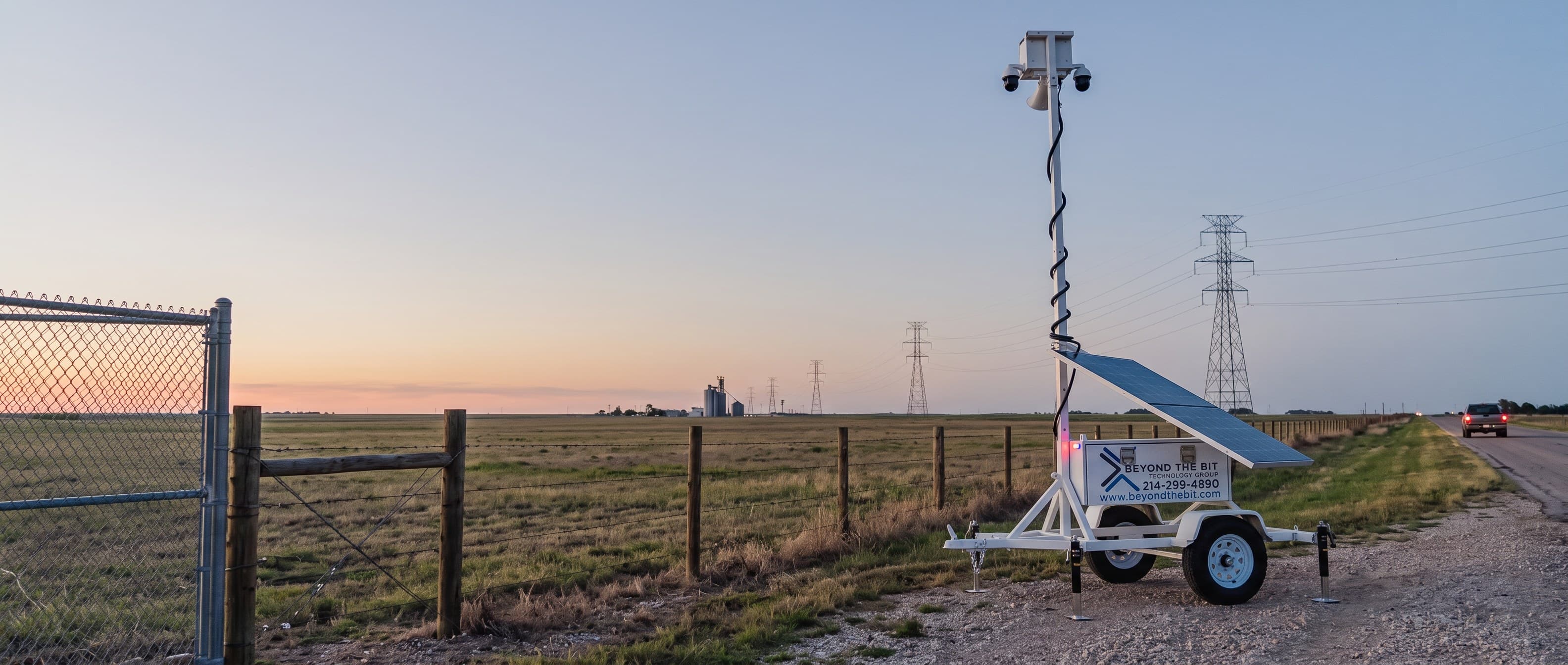 A Beyond the Bit security trailer on the gravel shoulder of a two-lane Texas highway at dusk, camera mast fully raised beside a ranch fence, transmission towers running to a flat horizon.