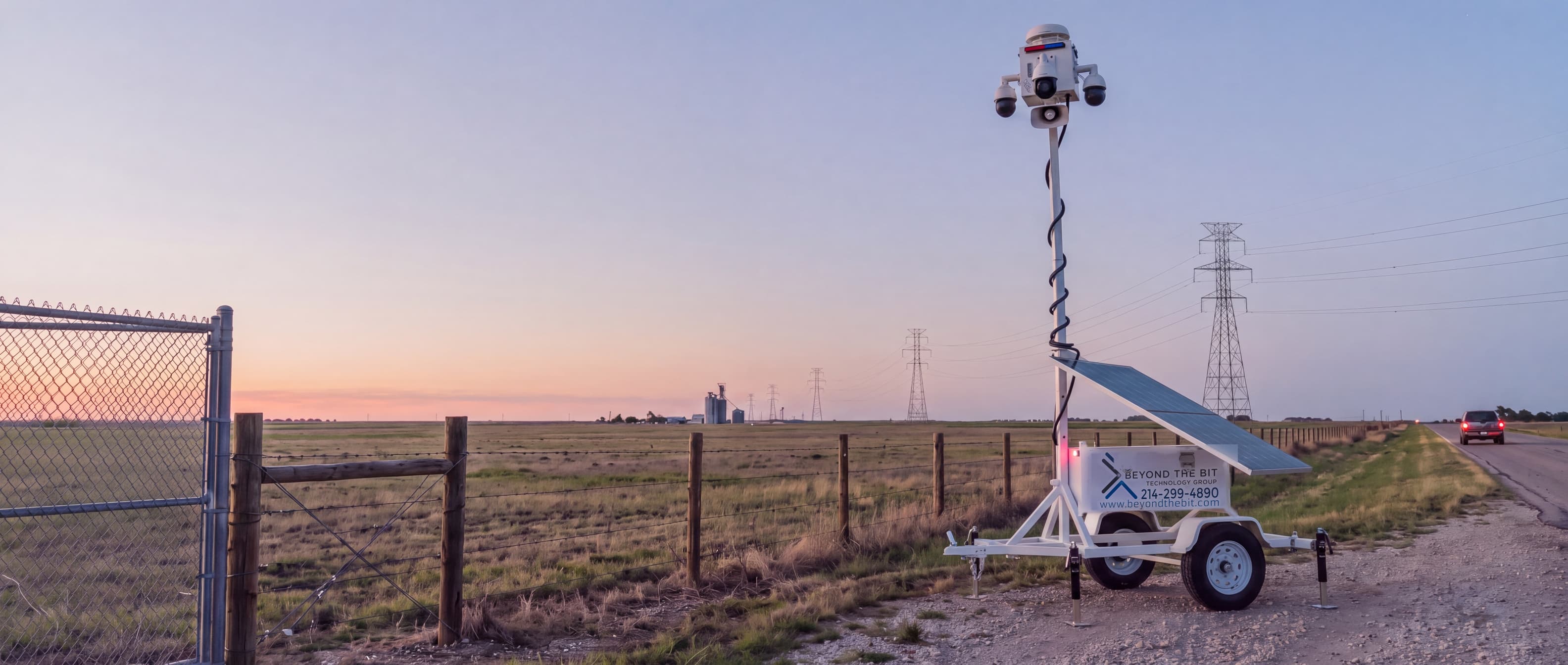 A Beyond the Bit security trailer on the gravel shoulder of a two-lane Texas highway at dusk, camera mast fully raised beside a ranch fence, transmission towers running to a flat horizon.