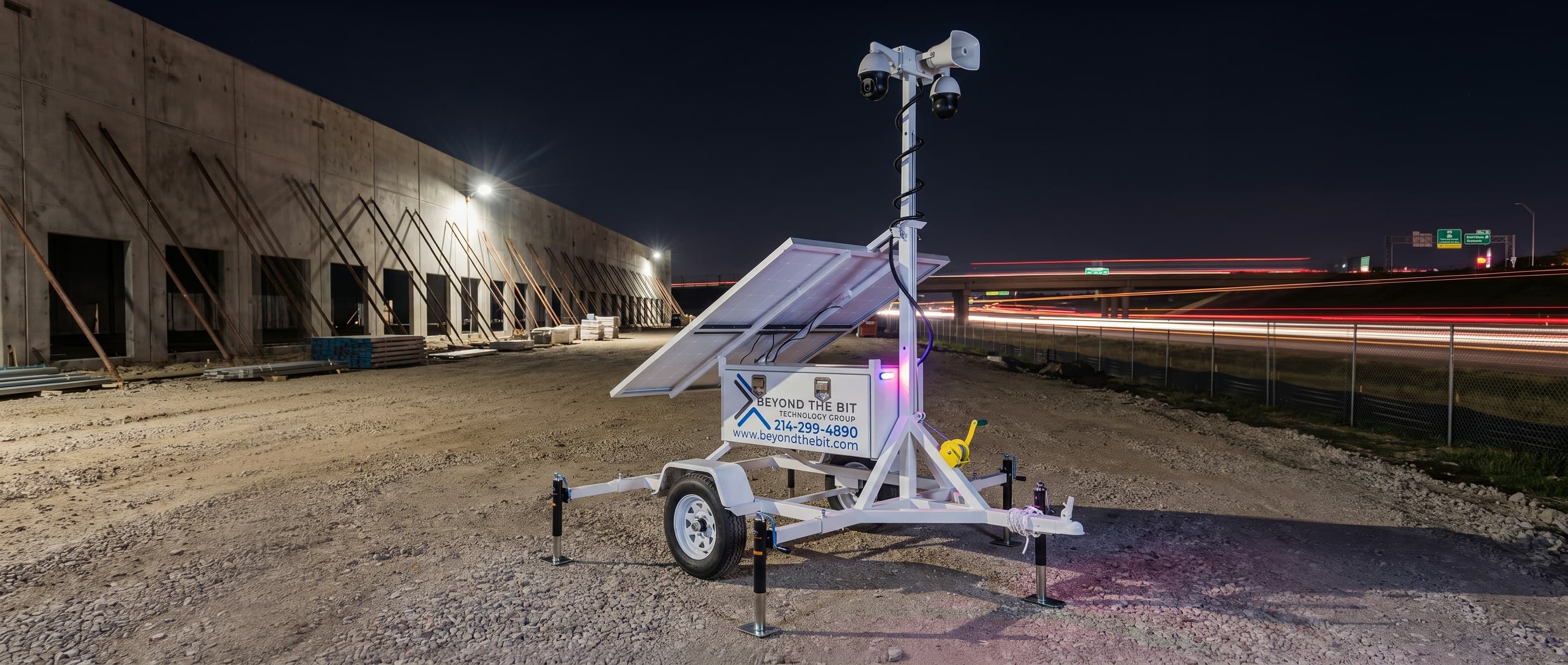 A Beyond the Bit security trailer at night in front of a braced tilt-wall warehouse shell, interstate traffic streaking past beyond the fence — representative of the I-35 corridor builds around Waco.