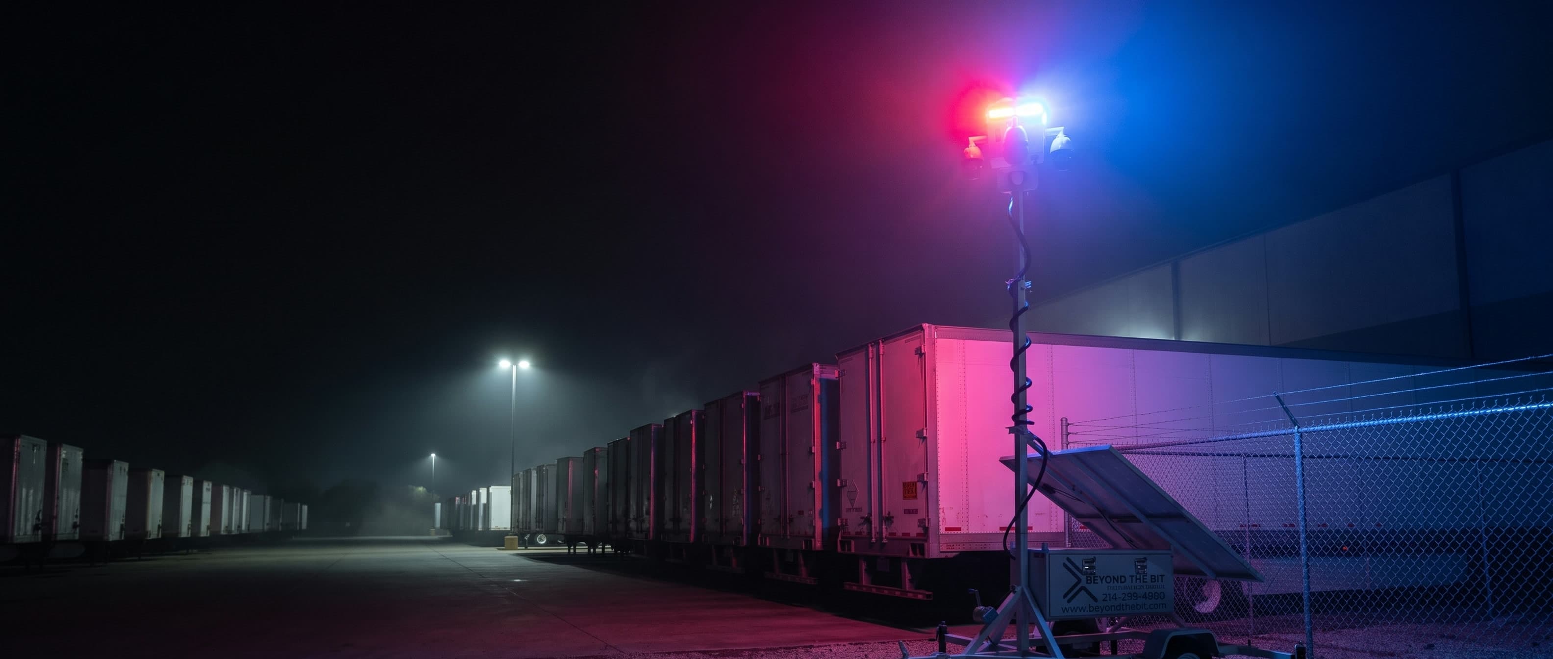 A Beyond the Bit security trailer at night in the corner of a distribution yard, camera mast fully raised and its red-and-blue strobe lit, a long row of parked semi trailers receding into ground haze beside it.