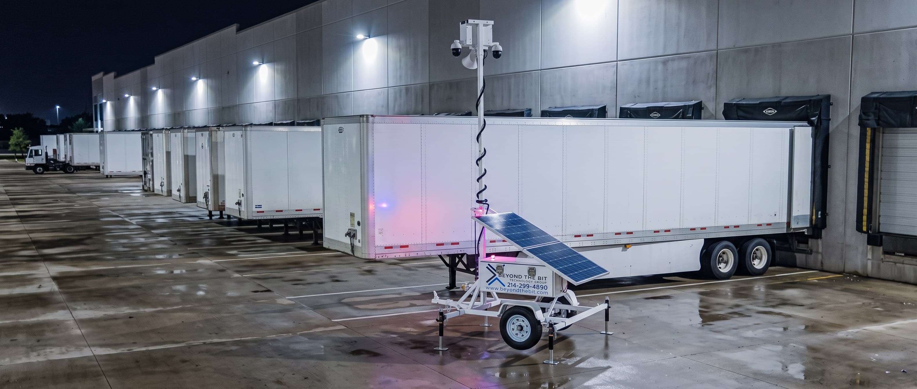 An elevated view of a Beyond the Bit security trailer on a wet warehouse truck court at night, its camera mast fully extended, a row of dry-van semi-trailers backed to the dock behind it under the building's wall lights.